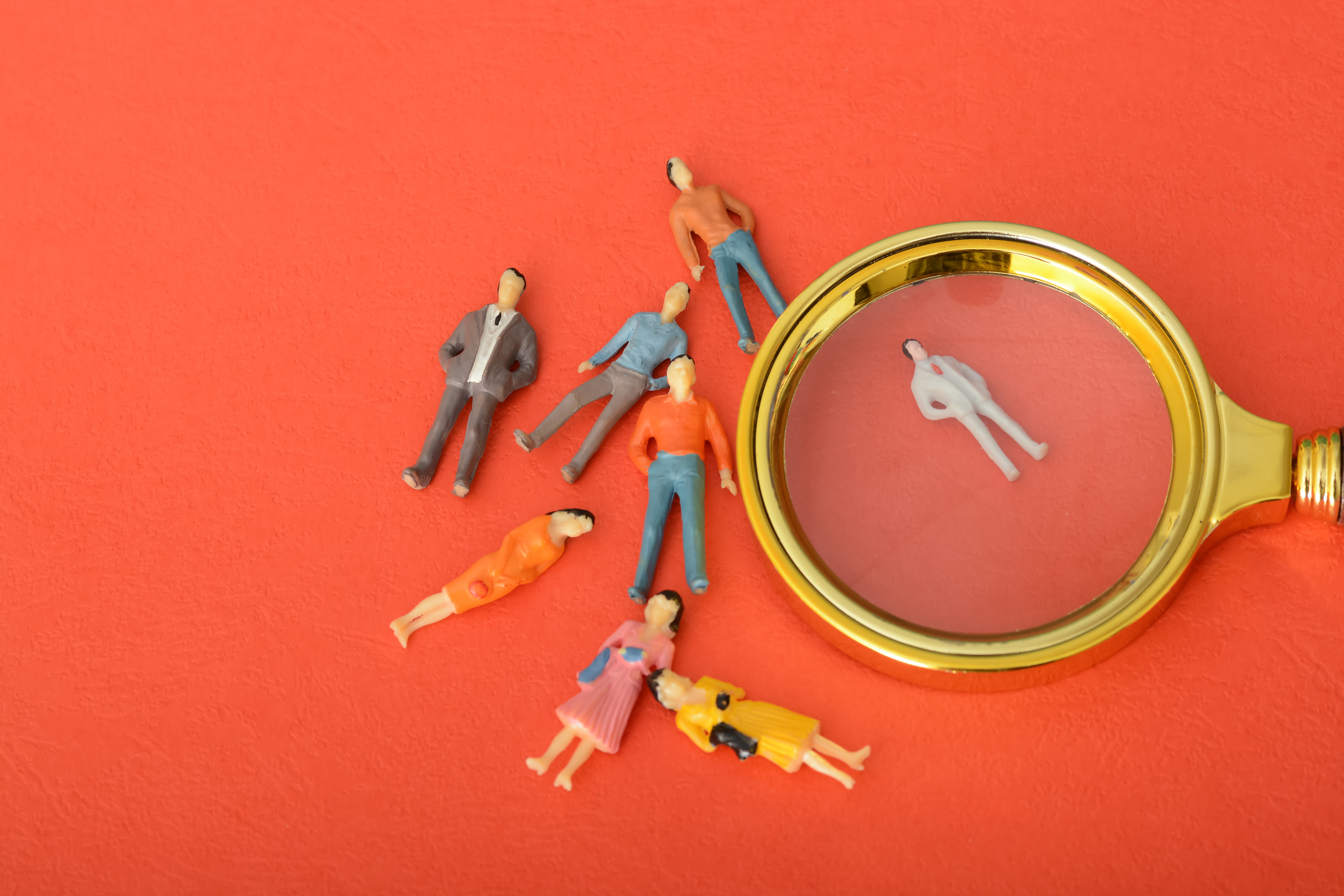 A magnifying glass over one person and missing a group of people outside it. It is all on a red background.